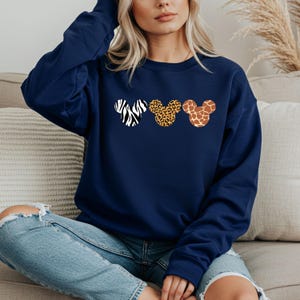 May include: Navy blue sweatshirt featuring three Mickey Mouse head silhouettes with animal print patterns: zebra, leopard, and giraffe. The sweatshirt is a long-sleeved top, perfect for casual wear.