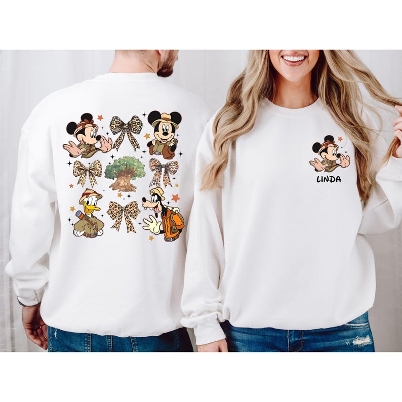 May include: Two white sweatshirts with a Disney theme. One sweatshirt features Mickey Mouse, Minnie Mouse, Donald Duck, and Goofy in safari outfits with bows and a tree. The other sweatshirt features Mickey Mouse in a safari outfit with the name "Linda" printed below.