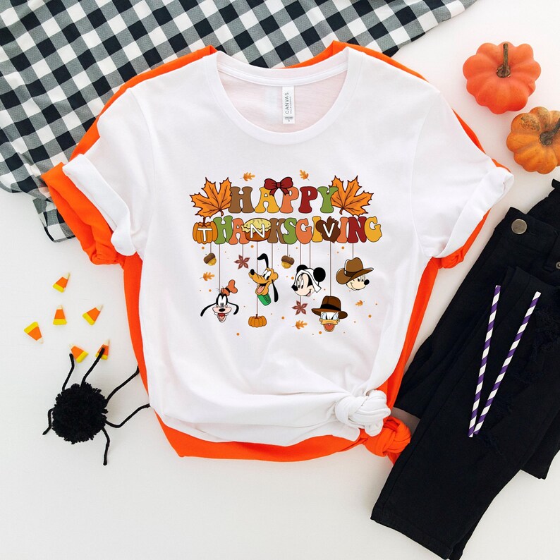 Op de afbeelding: Wit T-shirt met een kleurrijke "Happy Thanksgiving" afbeelding met stripfiguren en herfstbladeren. Het shirt wordt gecombineerd met een zwarte broek, pompoenen en Halloween-thema decoraties.