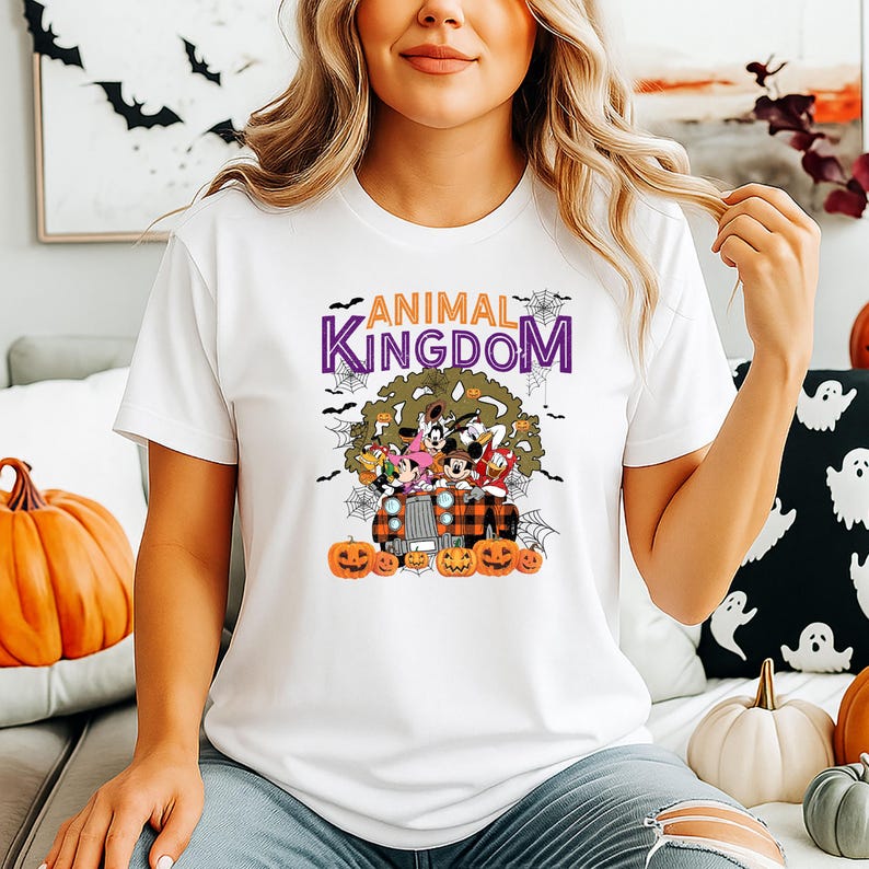 May include: White t-shirt with a colourful graphic of Disney characters on a truck with pumpkins. The text "ANIMAL KINGDOM" is above the graphic, with bats and spiderwebs. A fun top for Halloween or Disney enthusiasts.