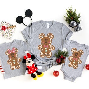 May include: Three grey t-shirts featuring a gingerbread man design with Mickey Mouse ears and a bow tie. The shirts are in different sizes, with the largest shirt having a red bow tie and the smallest shirt having a pink bow tie.