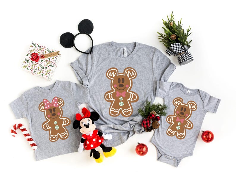 May include: Three grey t-shirts featuring a gingerbread man design with Mickey Mouse ears and a bow tie. The shirts are in different sizes, with the largest shirt having a red bow tie and the smallest shirt having a pink bow tie.