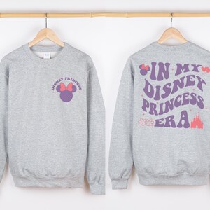 Puede incluir: Sudadera gris con un gr&aacute;fico de Disney Princess morado y rosa en la parte delantera y el texto "In My Disney Princess Era" en la parte posterior.