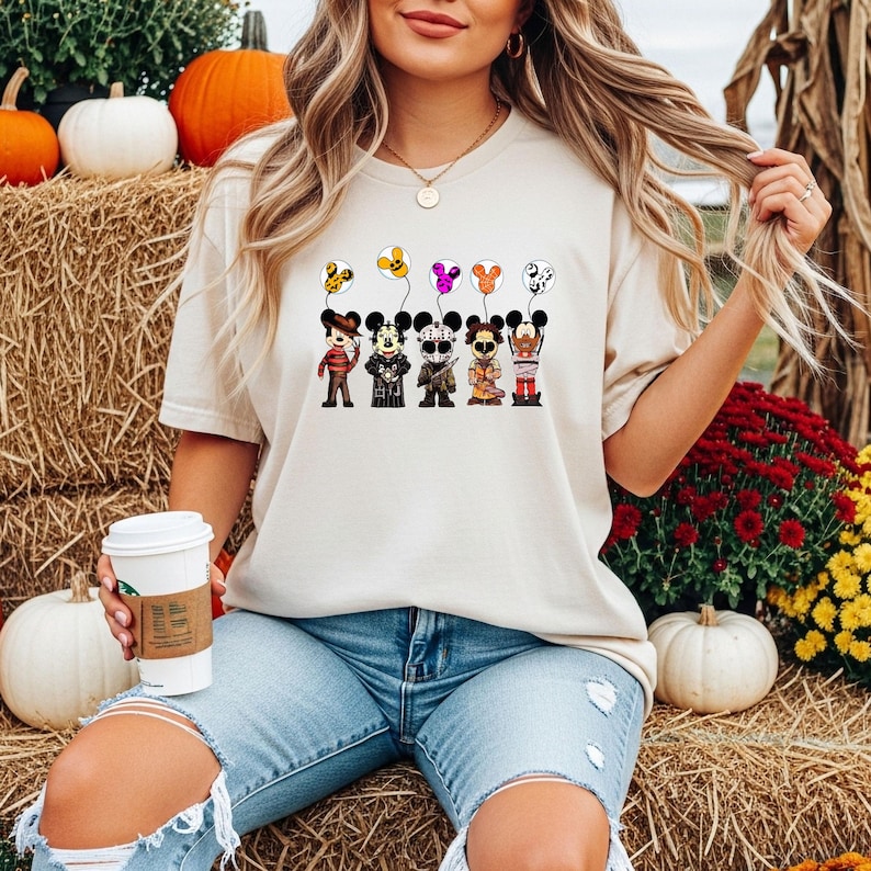 May include: Beige short-sleeved t-shirt featuring a graphic of cartoon characters dressed in Halloween costumes holding balloons. The balloons are various colors and patterns. The shirt is paired with blue jeans.