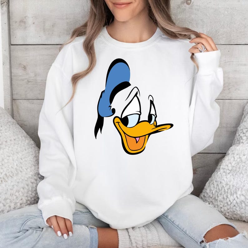 May include: White sweatshirt featuring a large graphic of Donald Duck's face. The cartoon character's face is in black, blue, yellow, and orange. The sweatshirt has long sleeves and a crew neck.