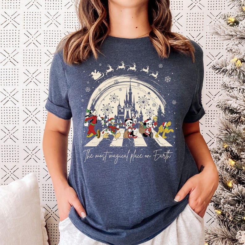 Puede incluir: Una camiseta azul marino con un dise&ntilde;o navide&ntilde;o que presenta a Mickey Mouse y sus amigos caminando por un paso de cebra frente al Castillo de Cenicienta. El texto "The most magical place on Earth" est&aacute; impreso debajo del dise&ntilde;o.