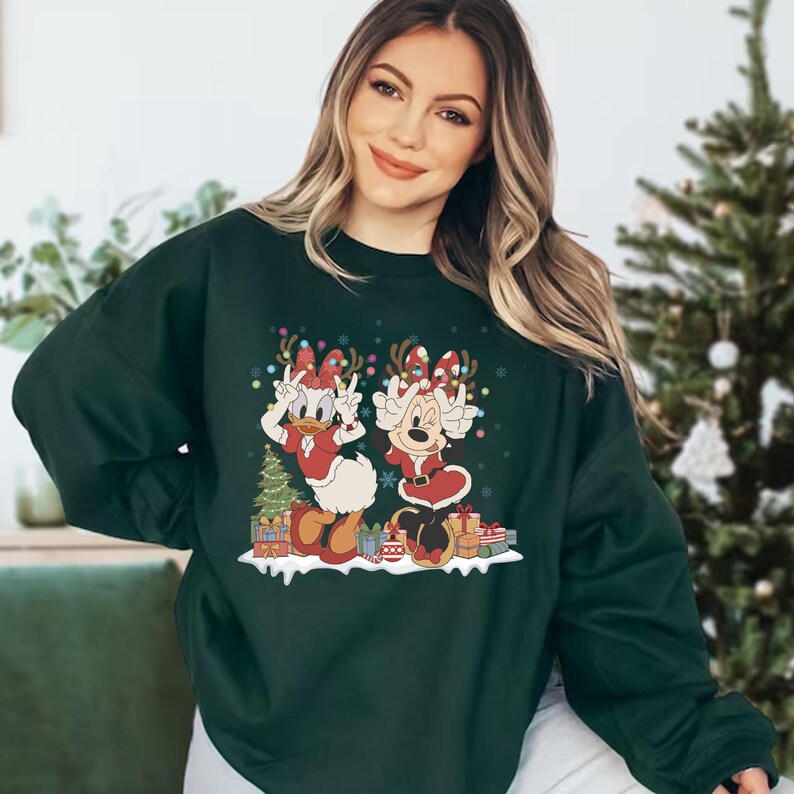 K&ouml;nnte beinhalten: Ein dunkelgr&uuml;ner Pullover mit einem weihnachtlichen Design, das Minnie Mouse und Daisy Duck zeigt, die Weihnachtsm&uuml;tzen tragen und Geschenke halten. Die Figuren sind von Schneeflocken und Weihnachtslichtern umgeben.