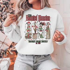 May include: White sweatshirt with a vintage-style graphic of Mickey Mouse and friends in front of the Haunted Mansion. The text reads "The Haunted Mansion Welcome Foolish Mortals Est. 1969".