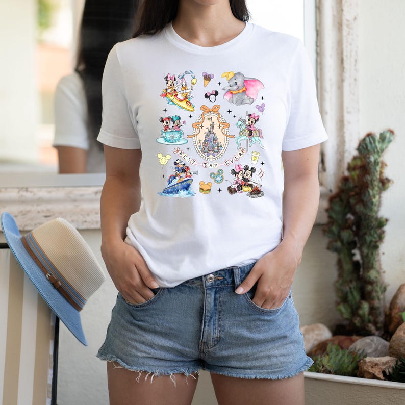 May include: White t-shirt with a colorful graphic print of various Disney characters, including Mickey Mouse, Minnie Mouse, Donald Duck, and Goofy. The graphic also includes the text "Best Day Ever".