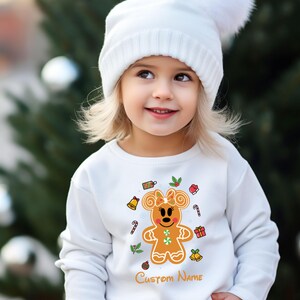 May include: White long-sleeved sweatshirt with a gingerbread Minnie Mouse design. The design includes a gingerbread Minnie Mouse with a red bow, surrounded by Christmas-themed elements like candy canes, bells, and gift boxes. The text "Custom Name" is printed below the design.