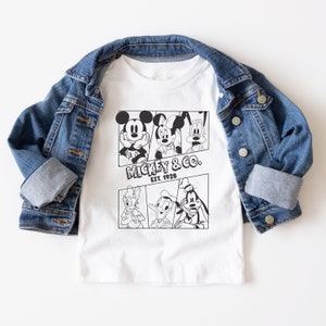 K&ouml;nnte beinhalten: Wei&szlig;es T-Shirt mit einem schwarz-wei&szlig;en Grafikdruck von Mickey Mouse, Minnie Mouse, Donald Duck und Goofy. Die Grafik zeigt die Figuren in Comic-Panels. Der Text "Mickey & Co. Est. 1928" ist unter der Grafik gedruckt.