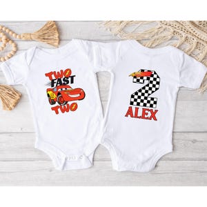 K&ouml;nnte beinhalten: Zwei wei&szlig;e Baby-Bodys. Einer zeigt ein rotes Rennauto mit dem Text "TWO FAST TWO". Der andere hat eine karierte Nummer 2 mit dem Namen "ALEX" in Rot. Ideal f&uuml;r den zweiten Geburtstag.