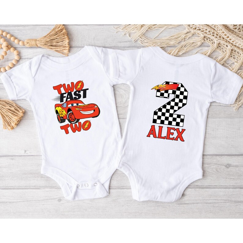 K&ouml;nnte beinhalten: Zwei wei&szlig;e Baby-Bodys. Einer zeigt ein rotes Rennauto mit dem Text "TWO FAST TWO". Der andere hat eine karierte Nummer 2 mit dem Namen "ALEX" in Rot. Ideal f&uuml;r den zweiten Geburtstag.