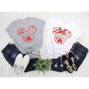 May include: Two t-shirts with a red Mickey Mouse head design featuring various Disney icons, including the castle, Minnie Mouse, and Mickey Mouse ears. The shirts are on a white surface with jeans and shoes.