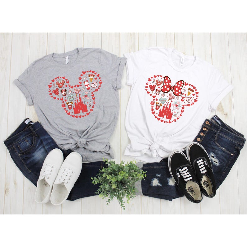 May include: Two t-shirts with a red Mickey Mouse head design featuring various Disney icons, including the castle, Minnie Mouse, and Mickey Mouse ears. The shirts are on a white surface with jeans and shoes.