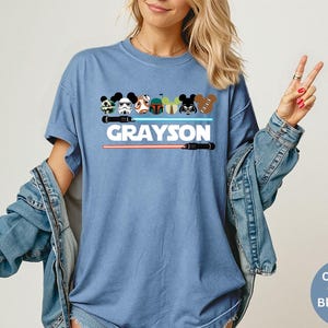 May include: Blue jean-colored t-shirt featuring cartoon character designs and the name "GRAYSON" in white block letters. Below the name are two lightsabers, one red and one black. The shirt has a relaxed fit.