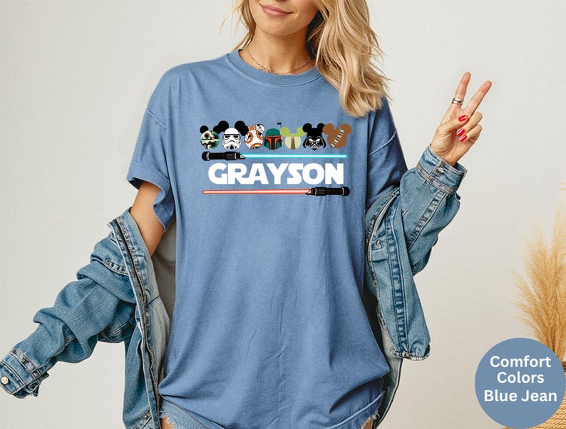 May include: Blue jean-colored t-shirt featuring cartoon character designs and the name "GRAYSON" in white block letters. Below the name are two lightsabers, one red and one black. The shirt has a relaxed fit.