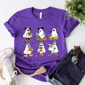 May include: Purple t-shirt featuring a graphic of Disney characters dressed as ghosts for Halloween. The characters include Mickey Mouse, Minnie Mouse, Donald Duck, Goofy, Pluto, and a ghost holding a pumpkin.