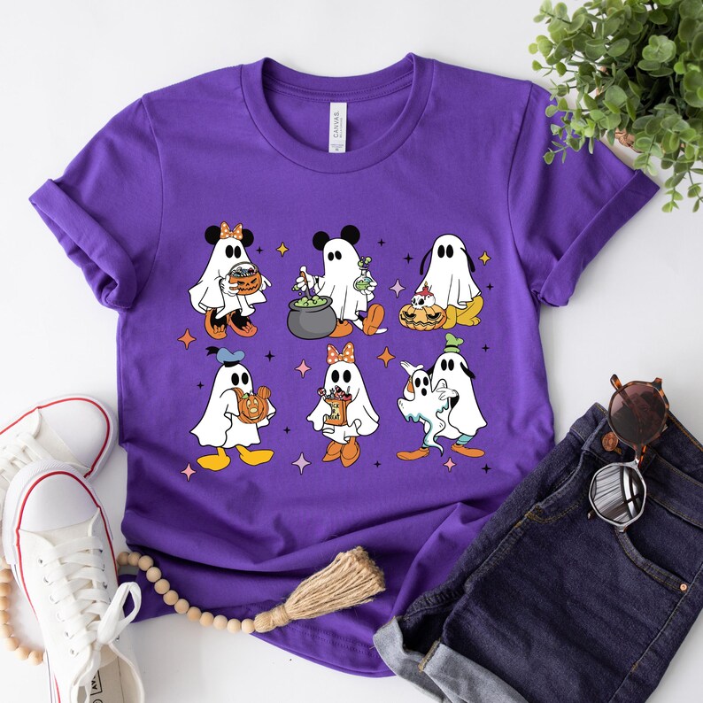May include: Purple t-shirt featuring a graphic of Disney characters dressed as ghosts for Halloween. The characters include Mickey Mouse, Minnie Mouse, Donald Duck, Goofy, Pluto, and a ghost holding a pumpkin.