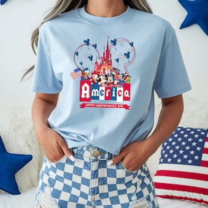 Może przedstawiać: Jasnoniebieska koszulka z patriotycznym wzorem z napisem "America" w kolorze czerwono-białym, postaciami z kresk&oacute;wek i zamkiem. Koszulka zawiera r&oacute;wnież napis "Happy Independence Day".