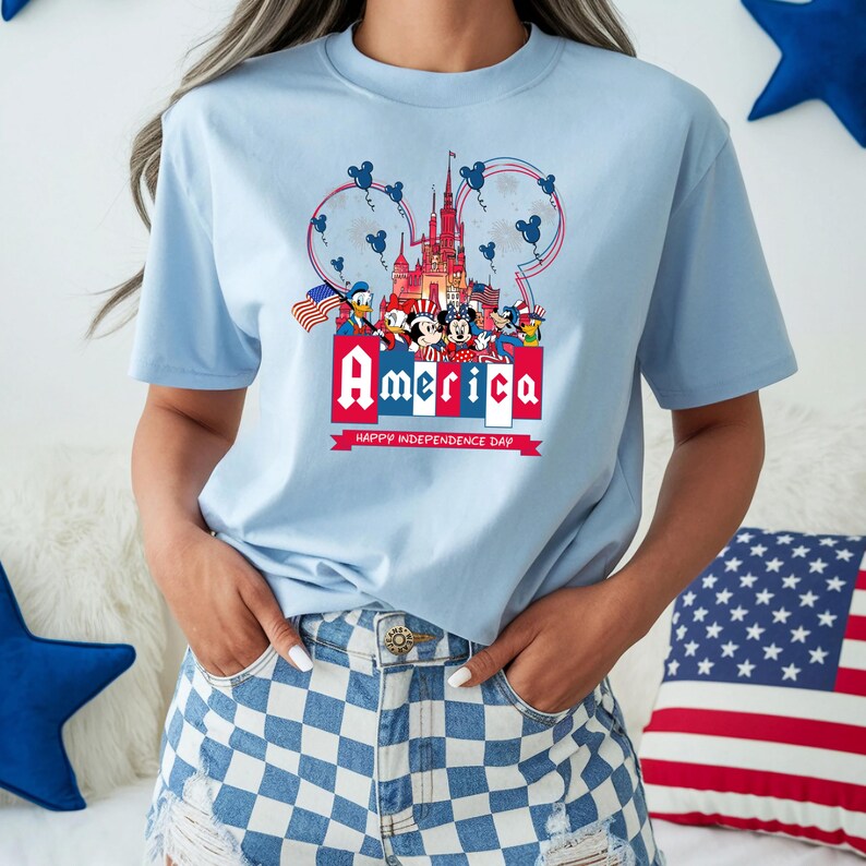 Może przedstawiać: Jasnoniebieska koszulka z patriotycznym wzorem z napisem "America" w kolorze czerwono-białym, postaciami z kresk&oacute;wek i zamkiem. Koszulka zawiera r&oacute;wnież napis "Happy Independence Day".