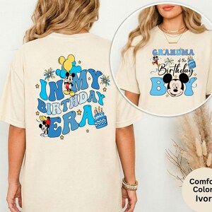 Op de afbeelding: Ivoor kleurig T-shirt met een blauw en zwart Mickey Mouse-ontwerp. Het shirt heeft de tekst "IN MY BIRTHDAY ERA" op de rug en "GRANDMA of the Birthday Boy" op de voorkant. Het ontwerp bevat sterren en een verjaardagstaart.