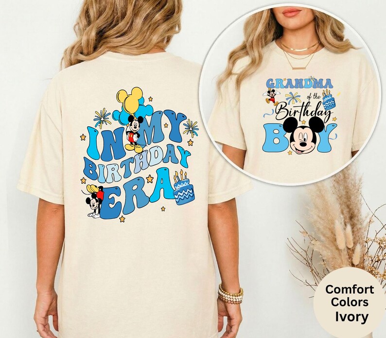 Op de afbeelding: Ivoor kleurig T-shirt met een blauw en zwart Mickey Mouse-ontwerp. Het shirt heeft de tekst "IN MY BIRTHDAY ERA" op de rug en "GRANDMA of the Birthday Boy" op de voorkant. Het ontwerp bevat sterren en een verjaardagstaart.