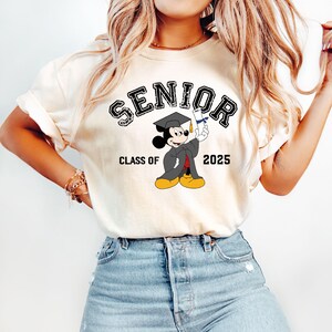 K&ouml;nnte beinhalten: Ein wei&szlig;es T-Shirt mit dem Text "SENIOR" in Schwarz mit einem Used-Look. Der Text befindet sich &uuml;ber einem Cartoon-Bild von Mickey Mouse, der eine Graduation Cap und eine Robe tr&auml;gt und ein Diplom h&auml;lt. Der Text "CLASS OF 2025" befindet sich unter dem Bild.
