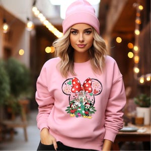 Puede incluir: Sudadera rosa con un dise&ntilde;o de Mickey Mouse y Minnie Mouse. El dise&ntilde;o muestra a Mickey y Minnie frente a un &aacute;rbol de Navidad con un castillo en el fondo. La sudadera tiene un lazo rosa en la parte superior de la cabeza.
