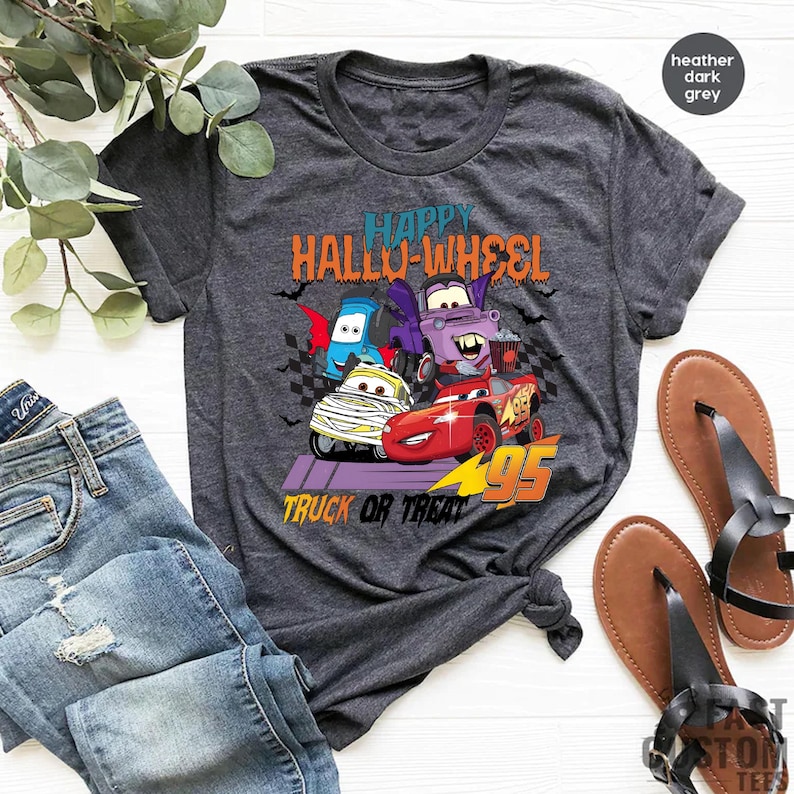 Może przedstawiać: Koszulka w kolorze ciemnoszary melanż z grafiką czterech kresk&oacute;wkowych samochod&oacute;w przebranych na Halloween. Na koszulce nadrukowany jest tekst "Happy Hallo-Wheel Truck or Treat".