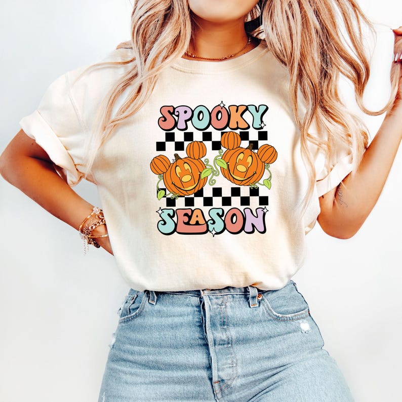 May include: Cream-colored t-shirt with a Halloween-themed graphic. The design features the words "Spooky Season" in colorful bubble letters, along with two smiling jack-o'-lanterns with Mickey Mouse ears on a black and white checkered background.