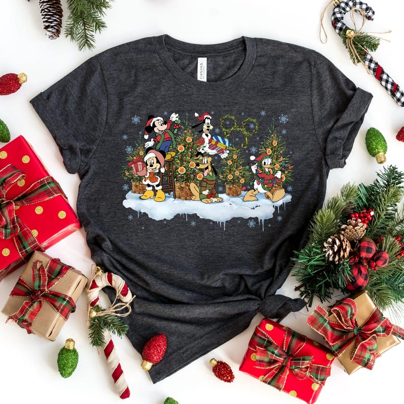 May include: A dark gray t-shirt with a Christmas-themed design featuring Mickey Mouse, Donald Duck, Goofy, and Pluto decorating a Christmas tree. The characters are wearing Santa hats and holding ornaments.