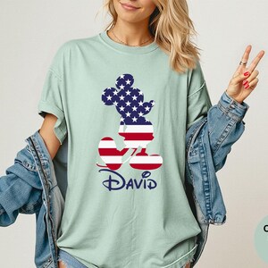 May include: A light green t-shirt featuring a Mickey Mouse silhouette filled with a US flag design. The name "David" is printed in navy blue script below the character. The shirt is part of the Comfort Colors Bay collection.