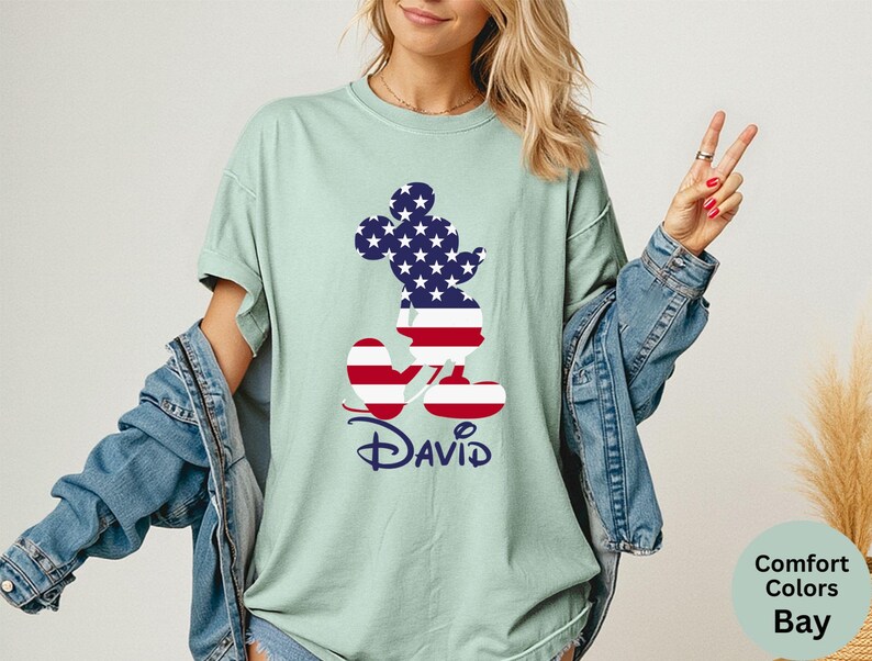 May include: A light green t-shirt featuring a Mickey Mouse silhouette filled with a US flag design. The name "David" is printed in navy blue script below the character. The shirt is part of the Comfort Colors Bay collection.