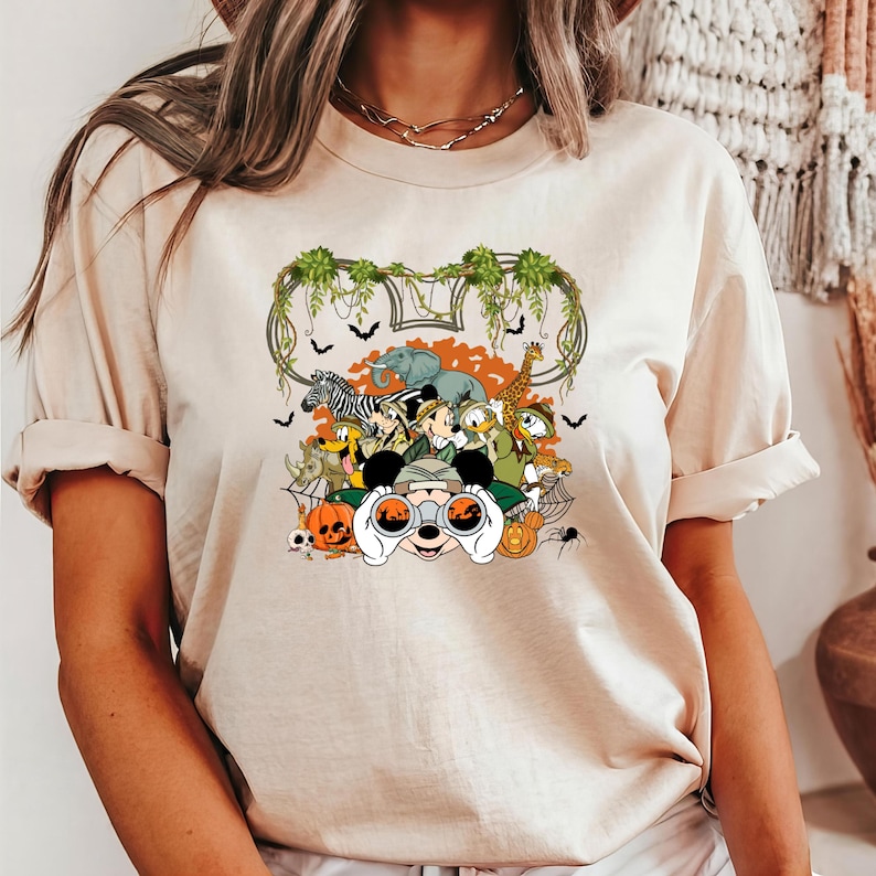 May include: Beige t-shirt featuring a colorful graphic of Mickey Mouse and friends in safari attire, surrounded by Halloween elements like pumpkins, bats, and a spiderweb. The design includes a Mickey Mouse-shaped frame with jungle foliage.