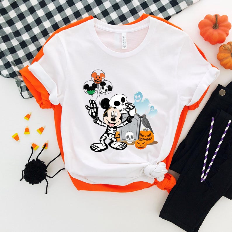 May include: White t-shirt with an orange trim featuring a Halloween-themed graphic. The graphic includes a skeleton-themed Mickey Mouse holding a skull and balloons, with pumpkins, a tombstone, and ghosts. The shirt is paired with black pants and Halloween decorations.