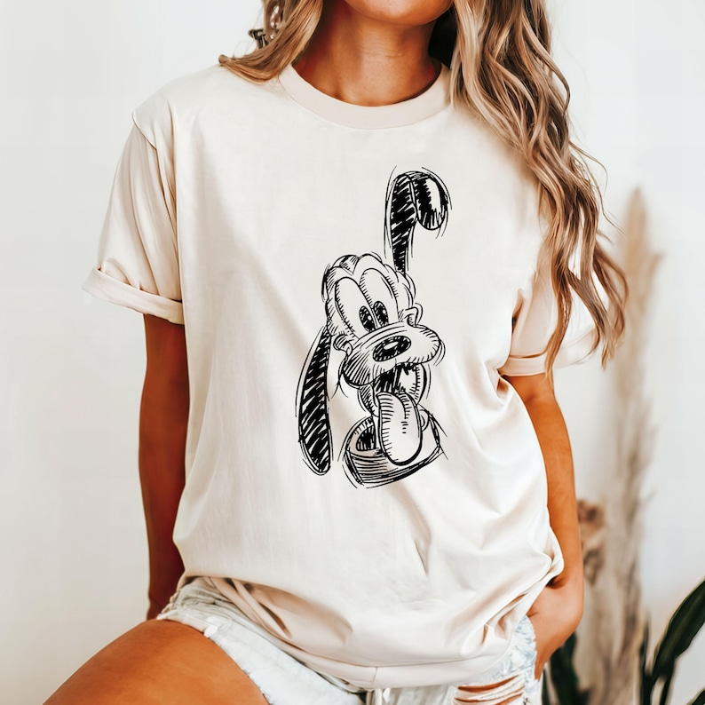 May include: A cream-colored t-shirt featuring a black and white sketch of Pluto, a cartoon dog. The graphic shows Pluto with his tongue out. The shirt has short sleeves and a crew neck.