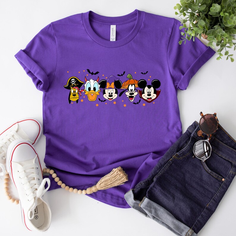Op de afbeelding: Paars T-shirt met een afbeelding van Mickey Mouse, Minnie Mouse, Donald Duck, Goofy en Pluto verkleed in Halloween-kostuums. De personages staan in een rij en hebben vleermuizen en pompoenen om zich heen.