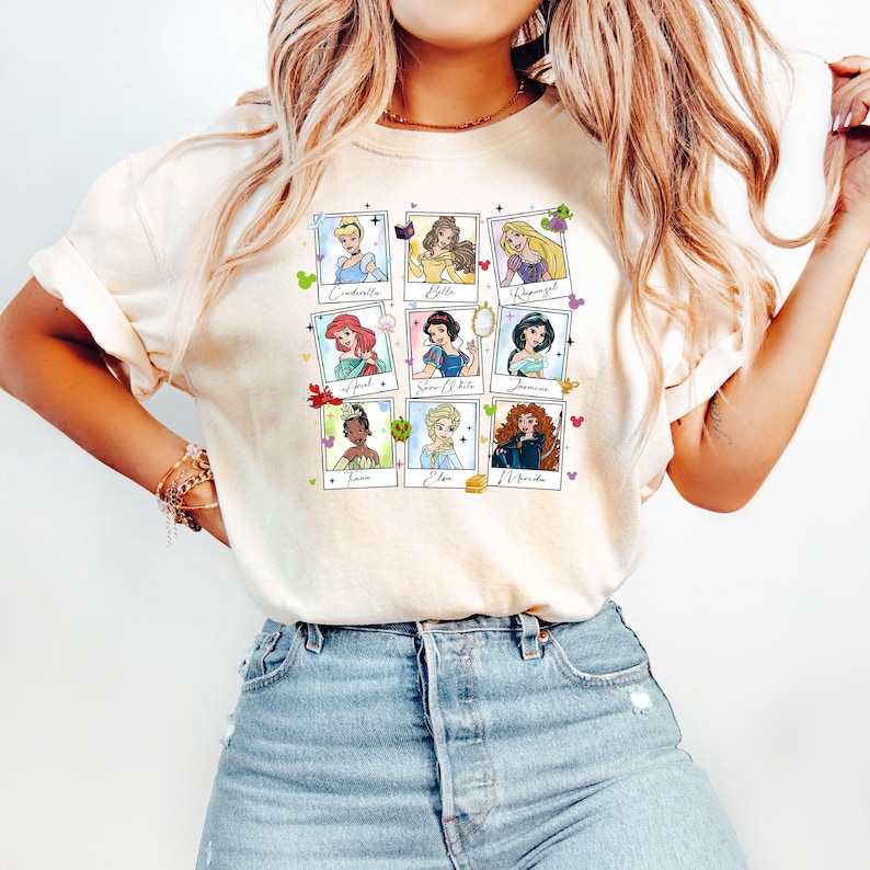 May include: Cream-colored t-shirt featuring a grid of nine Disney princess portraits in Polaroid-style frames. Each frame includes the princess's name. The shirt has short sleeves and a relaxed fit.