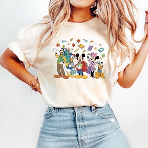 May include: Cream-colored t-shirt featuring a cartoon illustration of Disney characters, including Goofy, Donald Duck, Mickey Mouse, Minnie Mouse, and Pluto, with school supplies and autumn leaves. The characters are wearing backpacks and holding books.