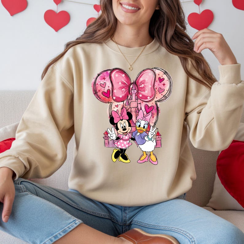 May include: Beige sweatshirt featuring a graphic of Minnie Mouse and Daisy Duck with a pink bow, castle, and hearts. The cartoon characters are in front of a pink castle. The sweatshirt has long sleeves and a crew neck.