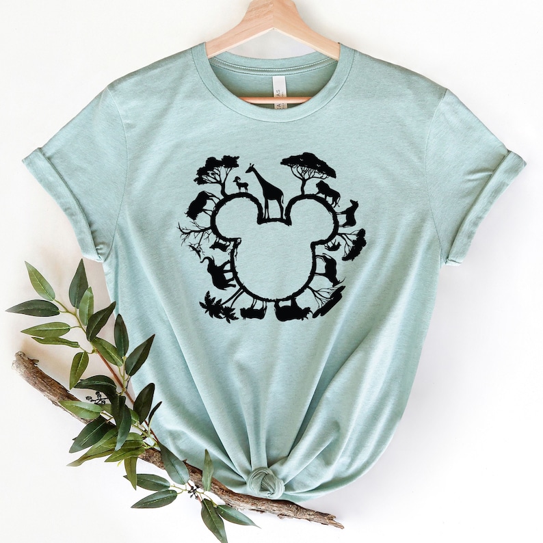 May include: A light blue t-shirt featuring a black silhouette of a Mickey Mouse head. The silhouette is formed by various animals, including a giraffe, zebra, and elephant, all surrounded by trees.