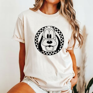 May include: A cream-coloured t-shirt with a black and white chequered circle graphic featuring a cartoon Goofy dog face.
