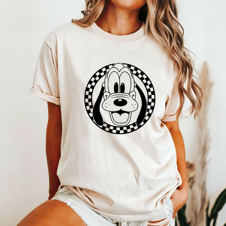 May include: A cream-coloured t-shirt with a black and white chequered circle graphic featuring a cartoon Goofy dog face.
