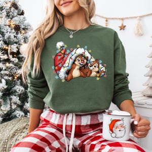 May include: Green sweatshirt featuring a festive graphic of Chip and Dale nestled in a Santa hat adorned with Christmas lights. The design is set against a green background. The person is wearing red and white plaid pajama pants and holding a Santa mug.