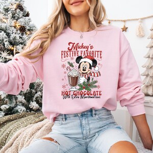 May include: Pink sweatshirt featuring a graphic of Mickey Mouse in a Santa hat and scarf, holding a cup of hot chocolate with marshmallows. The text reads "Mickey's Festive Favorite Hot Chocolate With Extra Marshmallows."