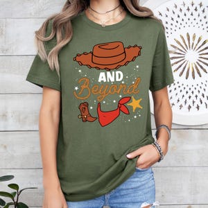 Peut inclure: T-shirt &agrave; manches courtes de couleur verte avec une image de dessin anim&eacute; d'un chapeau de cowboy, un bandana rouge, une botte de cowboy et une &eacute;toile. Le texte "AND BEYOND" est &eacute;crit dans une police de caract&egrave;res en forme de corde.