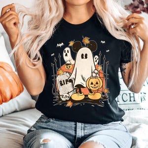 May include: Black t-shirt with a graphic of a ghost Mickey Mouse holding a jack-o'-lantern. The ghost is wearing Mickey Mouse ears and gloves. The graphic also includes a tombstone with the word "RIP" on it and other Halloween imagery.