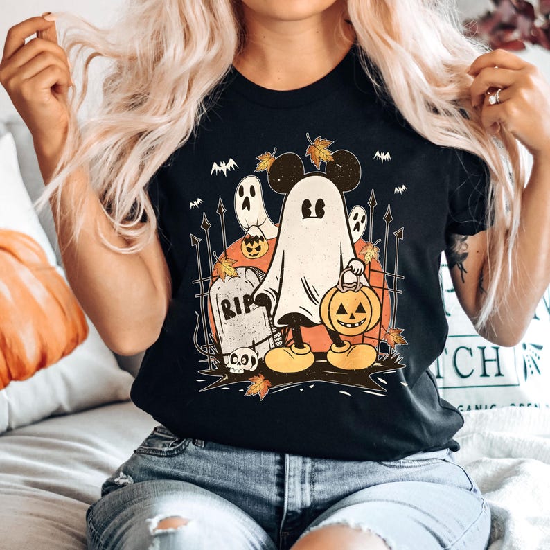 May include: Black t-shirt with a graphic of a ghost Mickey Mouse holding a jack-o'-lantern. The ghost is wearing Mickey Mouse ears and gloves. The graphic also includes a tombstone with the word "RIP" on it and other Halloween imagery.