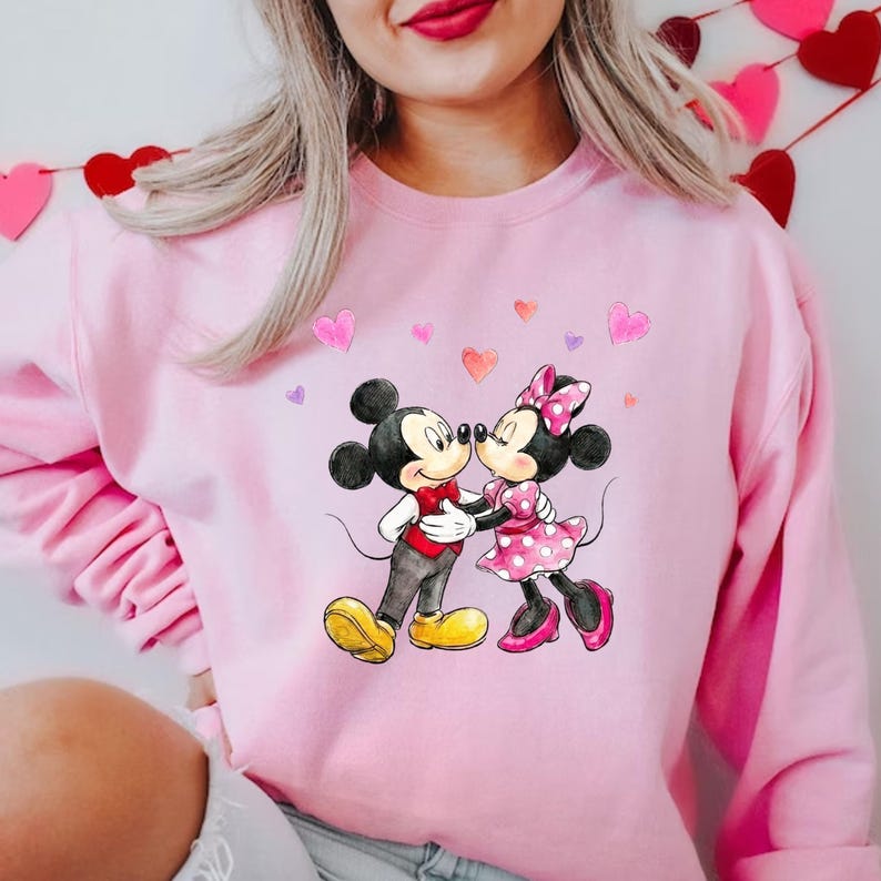 May include: A pale pink sweatshirt with a graphic of Mickey and Minnie Mouse in an embrace, with heart shapes around them. Mickey Mouse is in a red waistcoat and yellow shoes, Minnie Mouse wears a pink polka-dot dress and bow.